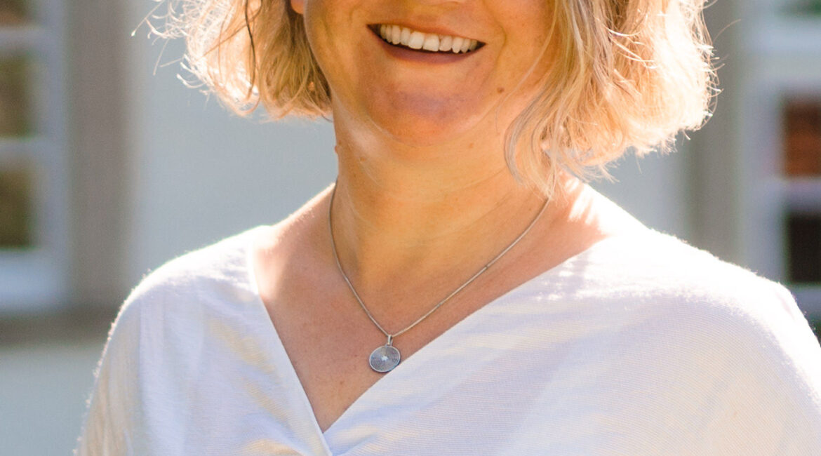 Eine lächelnde Frau mit kurzen, gewellten blonden Haaren, die ein weißes Oberteil und eine Halskette trägt, steht draußen im hellen Sonnenlicht, mit einem Gebäude und Fenstern im Hintergrund.