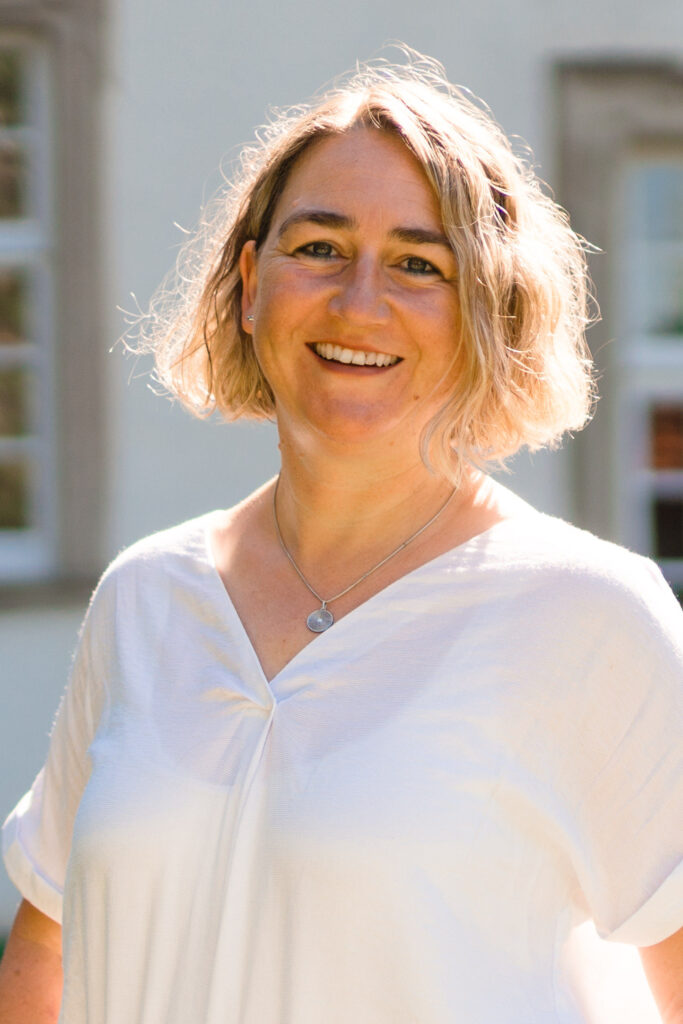 Eine lächelnde Frau mit kurzen, gewellten blonden Haaren, die ein weißes Oberteil und eine Halskette trägt, steht draußen im hellen Sonnenlicht, mit einem Gebäude und Fenstern im Hintergrund.