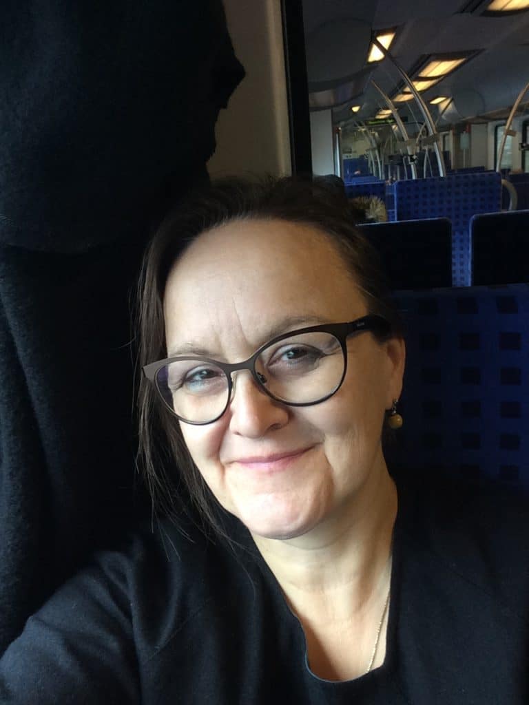 Eine Frau mit Brille und braunem Haar sitzt lächelnd auf einem blauen Sitz in einem Zug. Der Hintergrund zeigt leere Sitze und das Zuginnere, das sich im Fenster spiegelt.