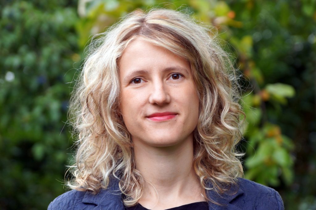 Eine Frau mit gewelltem blondem Haar und hellem Teint steht im Freien, trägt eine dunkle Jacke und lächelt leicht, während das grüne Laub im Hintergrund verschwimmt.