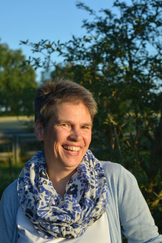 Eine Frau mit kurzen Haaren lächelt im Freien im Sonnenlicht, sie trägt eine hellblaue Strickjacke und einen blau-weiß gemusterten Schal. Im Hintergrund sind Bäume und Grünflächen zu sehen.