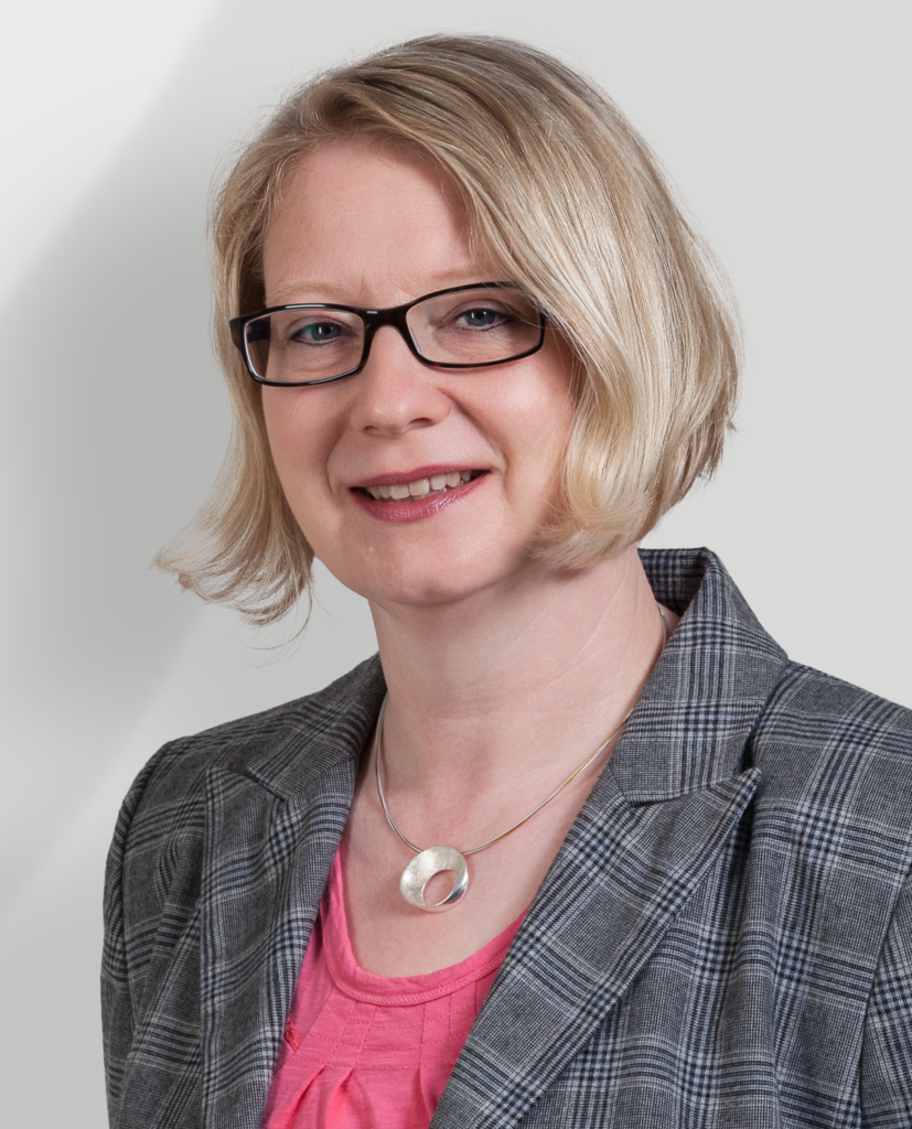 Eine Frau mit kurzen blonden Haaren und Brille, die einen grau karierten Blazer über einem rosafarbenen Oberteil und eine runde silberne Halskette trägt, lächelt vor einem hellen Hintergrund.