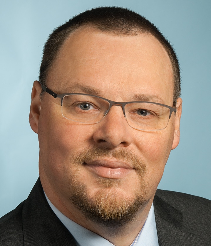 Ein Mann mit kurzen braunen Haaren, einem gestutzten Spitzbart und einer Brille, der einen dunklen Anzug und ein hellblaues Hemd trägt, lächelt leicht vor einem einfarbigen hellblauen Hintergrund.