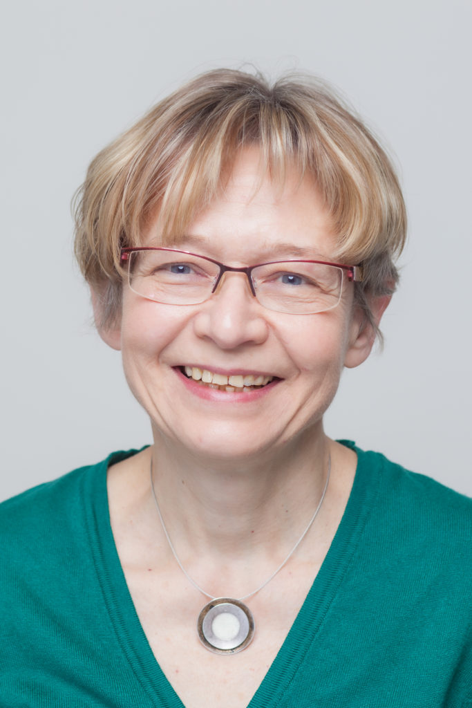 Eine lächelnde ältere Frau mit kurzen, hellen Haaren, die eine Brille, ein grünes Oberteil und eine runde Halskette mit Anhänger trägt, posiert vor einem schlichten hellen Hintergrund.