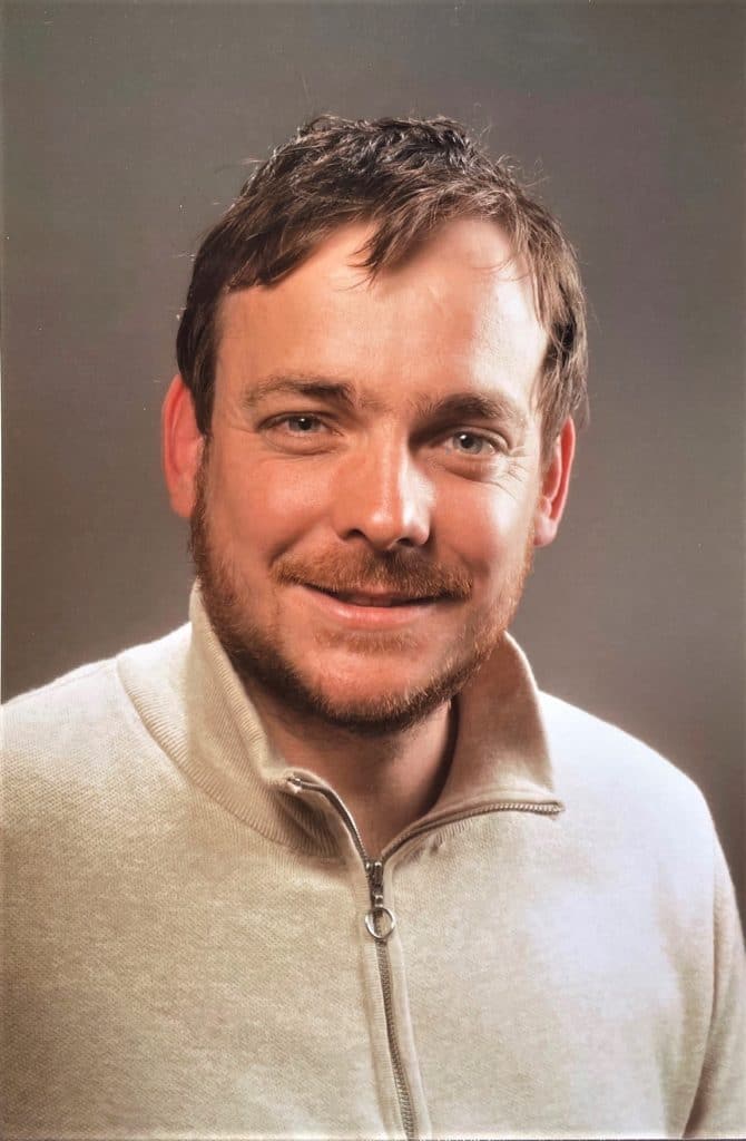 Ein Mann mit kurzen braunen Haaren und Bart, lächelnd, trägt einen hellbeigen Pullover mit Reißverschluss und posiert vor einem schlichten grauen Hintergrund.