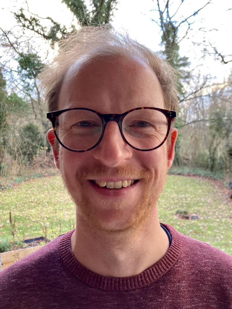 Eine lächelnde Person mit kurzen hellen Haaren und Brille, die einen kastanienbraunen Pullover trägt, steht draußen vor einem grasbewachsenen Hof mit Bäumen und heruntergefallenen Blättern im Hintergrund.