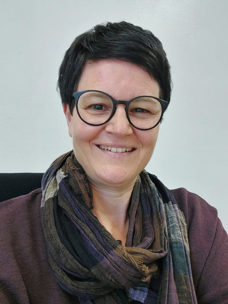 Eine Frau mit kurzen dunklen Haaren und einer runden Brille lächelt in die Kamera. Sie trägt einen dunklen, mehrfarbigen Schal und ein braunes Oberteil vor einem schlichten hellen Hintergrund.