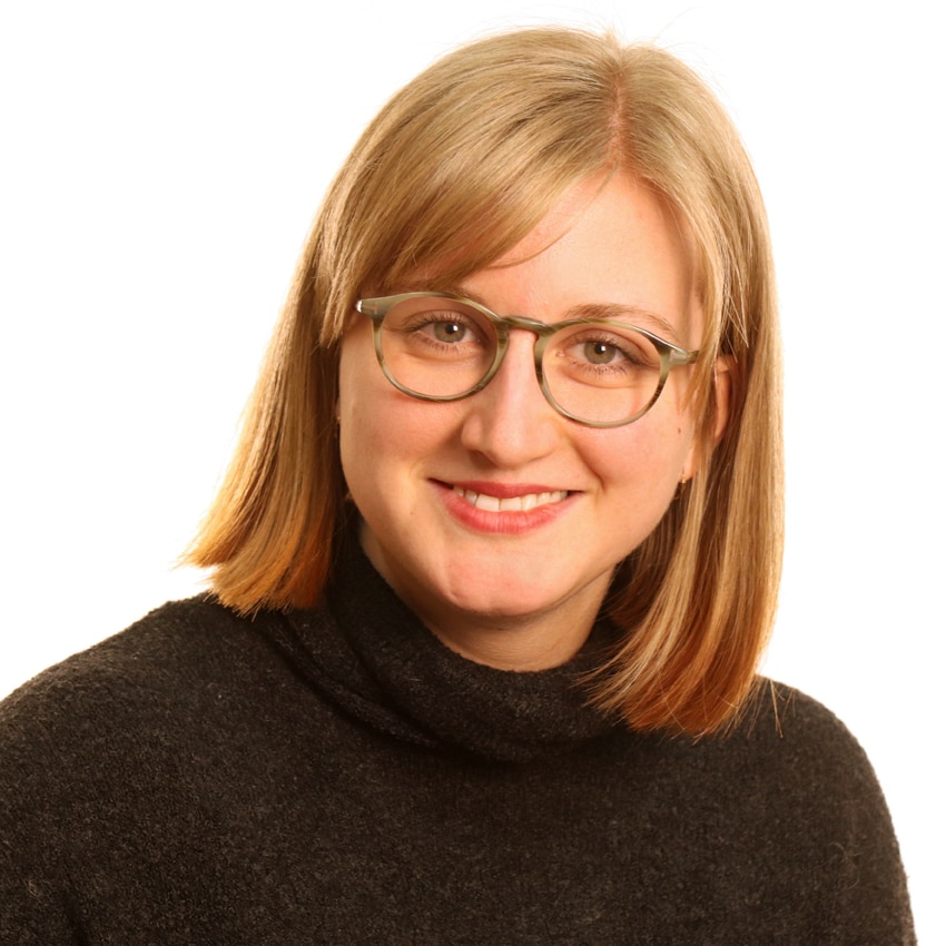 Eine Frau mit glattem, schulterlangem blondem Haar und Brille lächelt in die Kamera. Sie trägt einen dunklen Rollkragenpullover und ist vor einem schlichten weißen Hintergrund abgebildet.