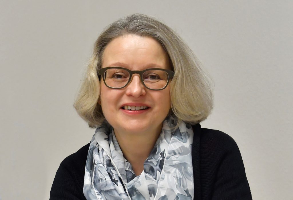 Eine Frau mit schulterlangen grauen Haaren, die eine Brille, ein schwarzes Oberteil und einen gemusterten Schal trägt, lächelt, während sie vor einem schlichten hellen Hintergrund sitzt.