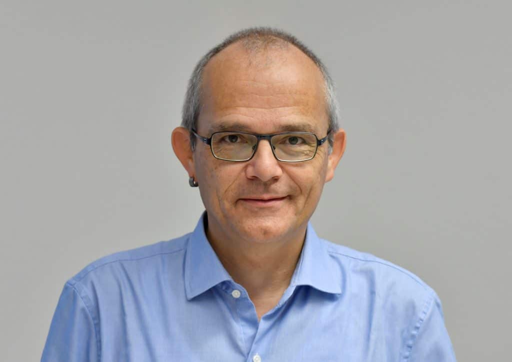 Ein Mann mittleren Alters mit kurzen grauen Haaren, einer Brille und einem Ohrring trägt ein hellblaues Button-up-Hemd und lächelt sanft vor einem schlichten hellgrauen Hintergrund.
