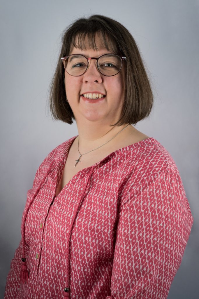 Eine Frau mit kinnlangem braunem Haar und Brille lächelt in die Kamera. Sie trägt eine gemusterte rosa Bluse und eine Halskette und steht vor einem schlichten hellgrauen Hintergrund.