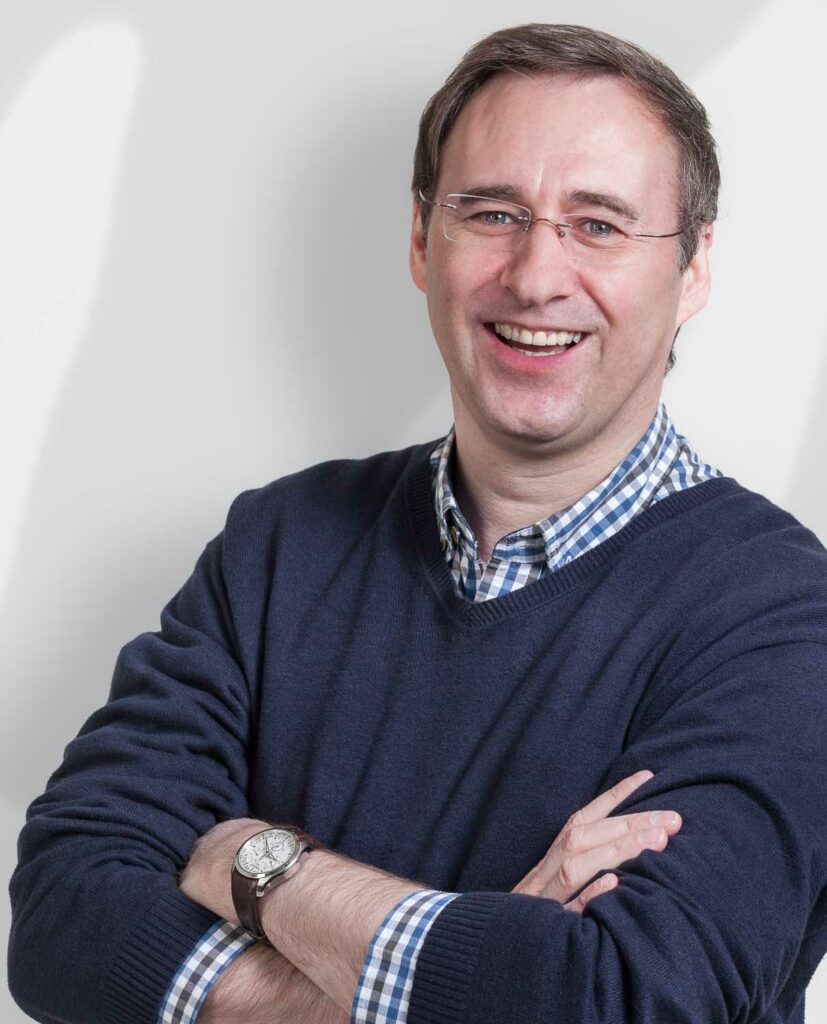 Ein Mann mit kurzen braunen Haaren, der eine Brille, einen marineblauen Pullover und ein kariertes Hemd trägt, steht mit verschränkten Armen und lächelt in die Kamera vor einem hellen Hintergrund.