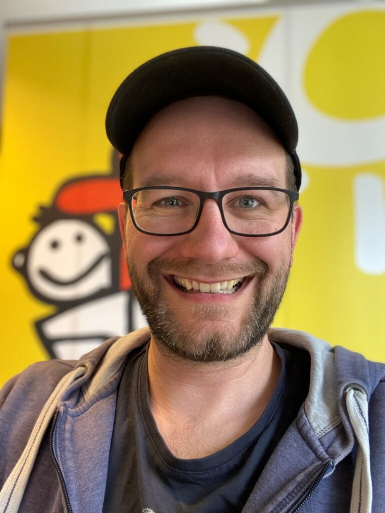 Ein lächelnder Mann mit Brille und Bart trägt eine schwarze Mütze und einen blauen Kapuzenpulli und posiert vor einem leuchtend gelben Hintergrund mit einer fröhlichen Zeichentrickfigur.