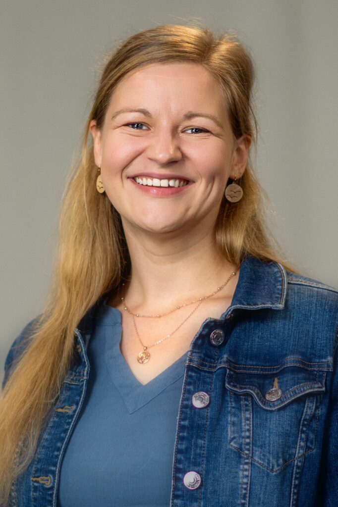 Eine Frau mit langen blonden Haaren, die ein blaues Hemd, eine Jeansjacke und Goldschmuck trägt, lächelt in die Kamera vor einem schlichten, hellen Hintergrund.
