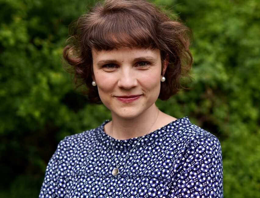 Eine Frau mit kurzen braunen Haaren, die ein gemustertes blaues Oberteil und Perlenohrringe trägt, steht mit verschränkten Armen im Freien und lächelt sanft vor einem üppigen grünen Laub.