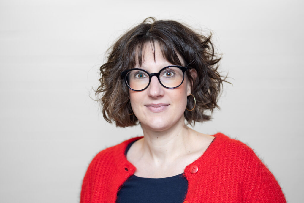 Frau mit gewelltem braunem Haar, die eine schwarze Brille, einen roten Pullover und ein dunkles Hemd trägt, steht vor einem hellen Hintergrund.