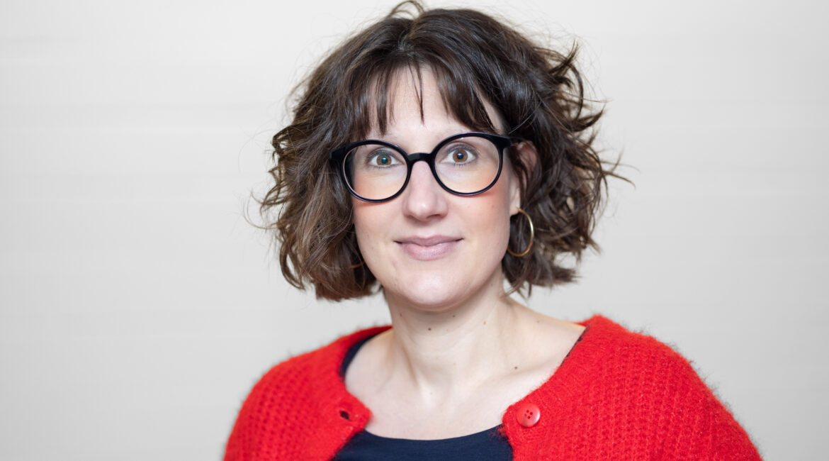 Frau mit gewelltem braunem Haar, die eine schwarze Brille, einen roten Pullover und ein dunkles Hemd trägt, steht vor einem hellen Hintergrund.