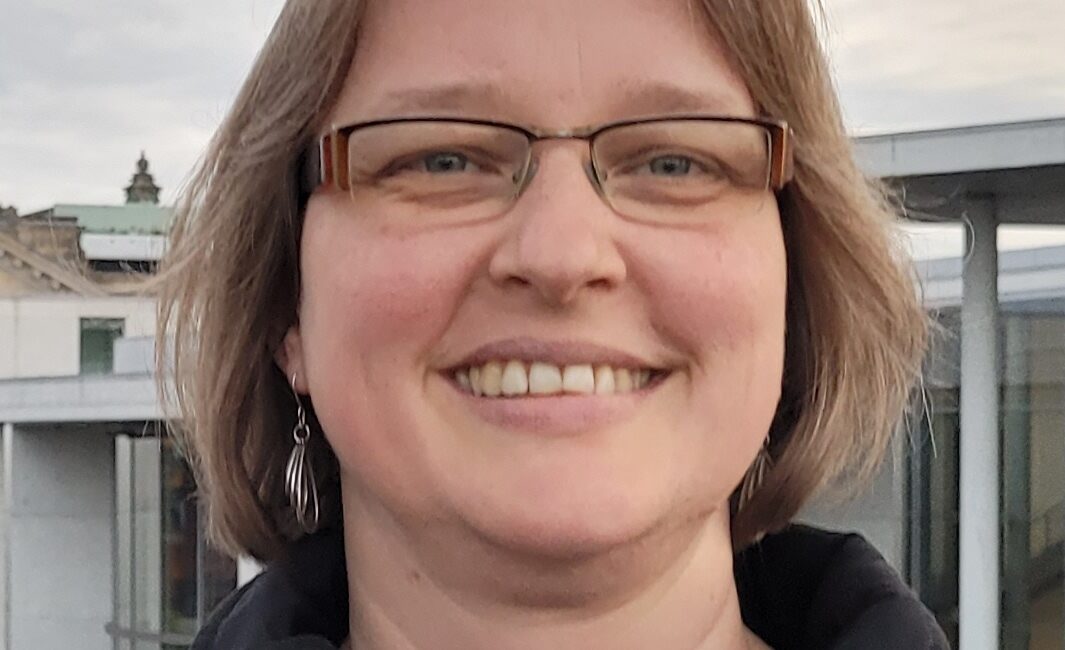 Eine Person mit kurzen hellbraunen Haaren und Brille, lächelnd und mit einer dunklen Jacke bekleidet, steht im Freien mit Gebäuden und einem bewölkten Himmel im Hintergrund.
