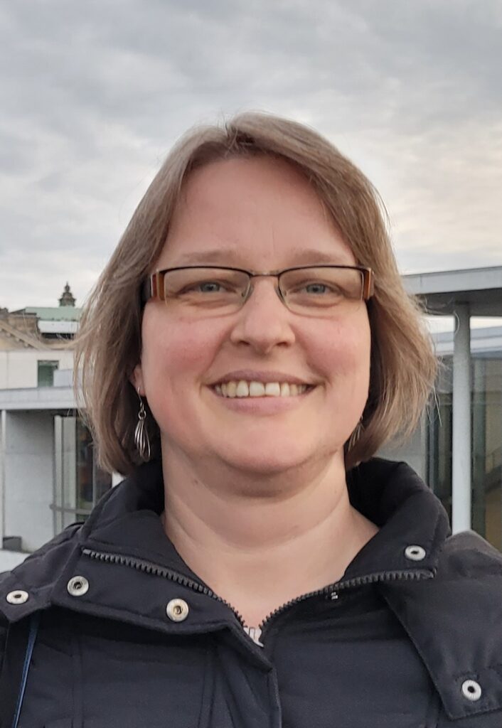 Eine Person mit kurzen hellbraunen Haaren und Brille, lächelnd und mit einer dunklen Jacke bekleidet, steht im Freien mit Gebäuden und einem bewölkten Himmel im Hintergrund.