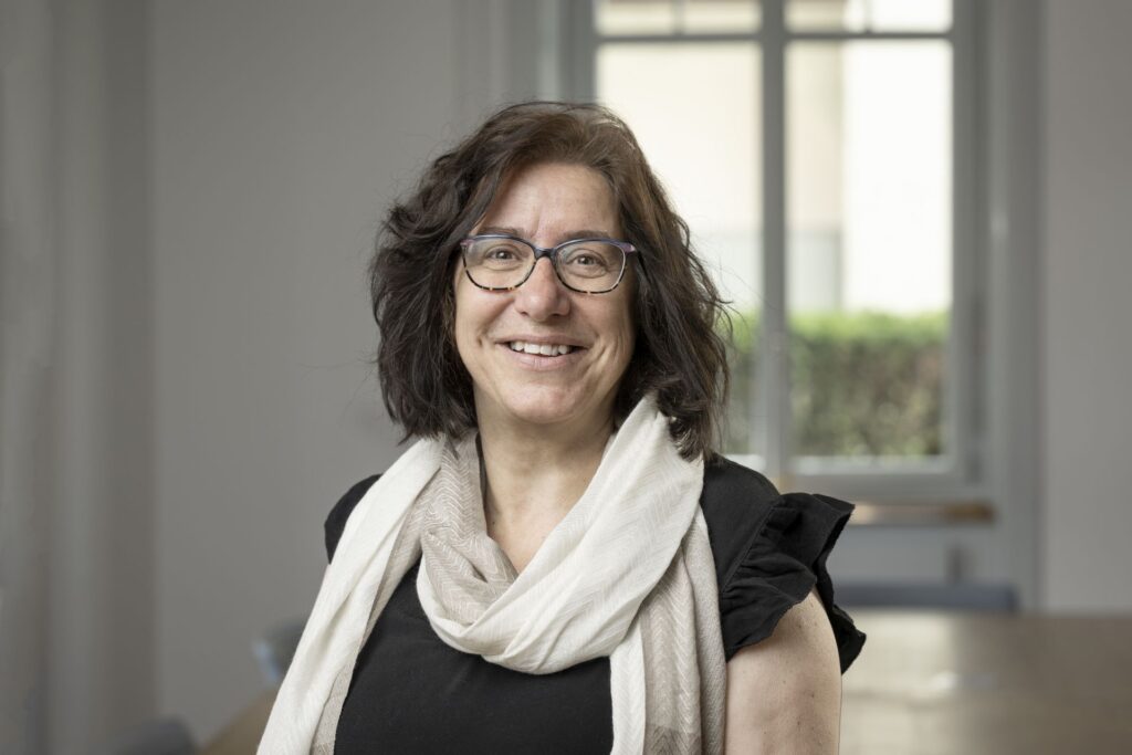 Eine Frau mit schulterlangem braunem Haar und Brille lächelt, sie trägt einen hellen Schal und ein schwarzes Oberteil und steht in einem hellen Raum mit einem Fenster im Hintergrund.