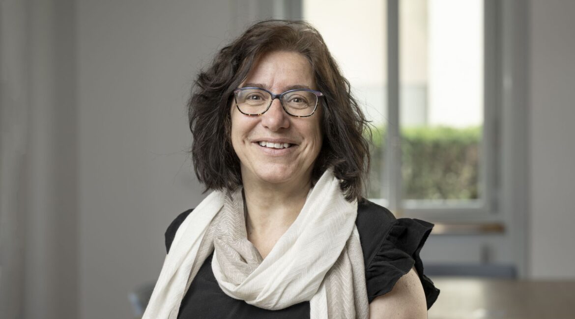 Eine Frau mit schulterlangem braunem Haar und Brille lächelt, sie trägt einen hellen Schal und ein schwarzes Oberteil und steht in einem hellen Raum mit einem Fenster im Hintergrund.