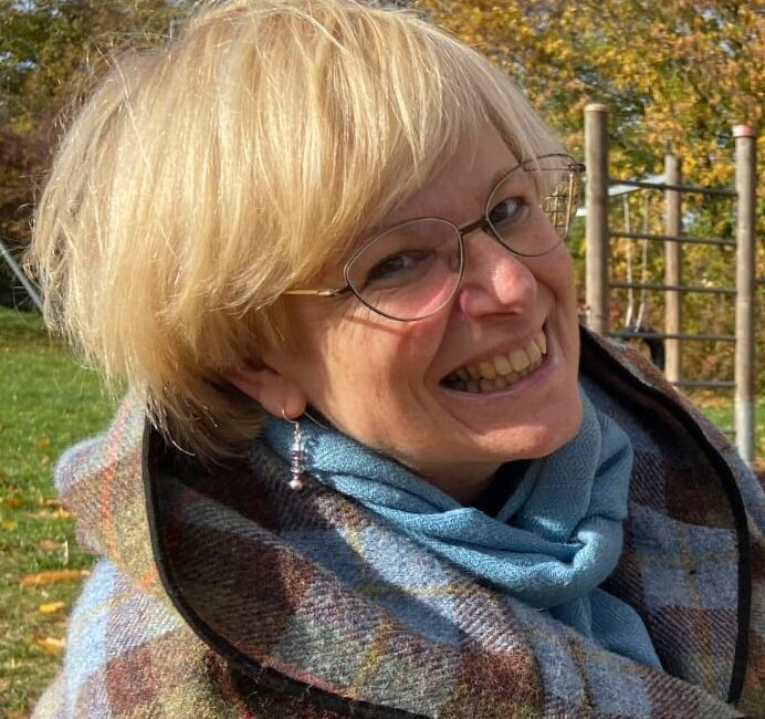 Eine lächelnde Frau mit kurzen blonden Haaren, einer Brille und einem blauen Schal steht an einem sonnigen Tag in einem Park, eingewickelt in einen karierten Mantel.