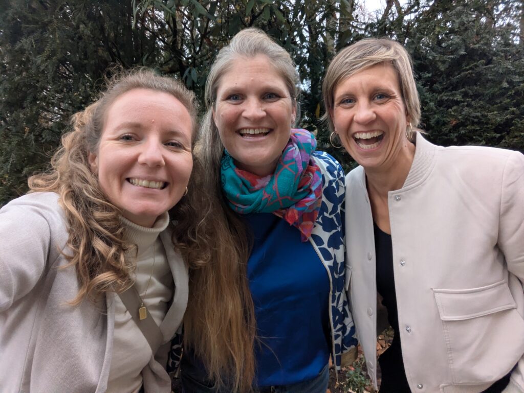 Drei Frauen lächeln und posieren zusammen für ein Selfie im Freien, mit Bäumen und Grün im Hintergrund.