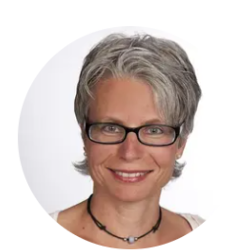 Lächelnde Frau mit kurzen grauen Haaren, die eine Brille und eine schwarze Perlenkette trägt, vor einem schlichten weißen Hintergrund.