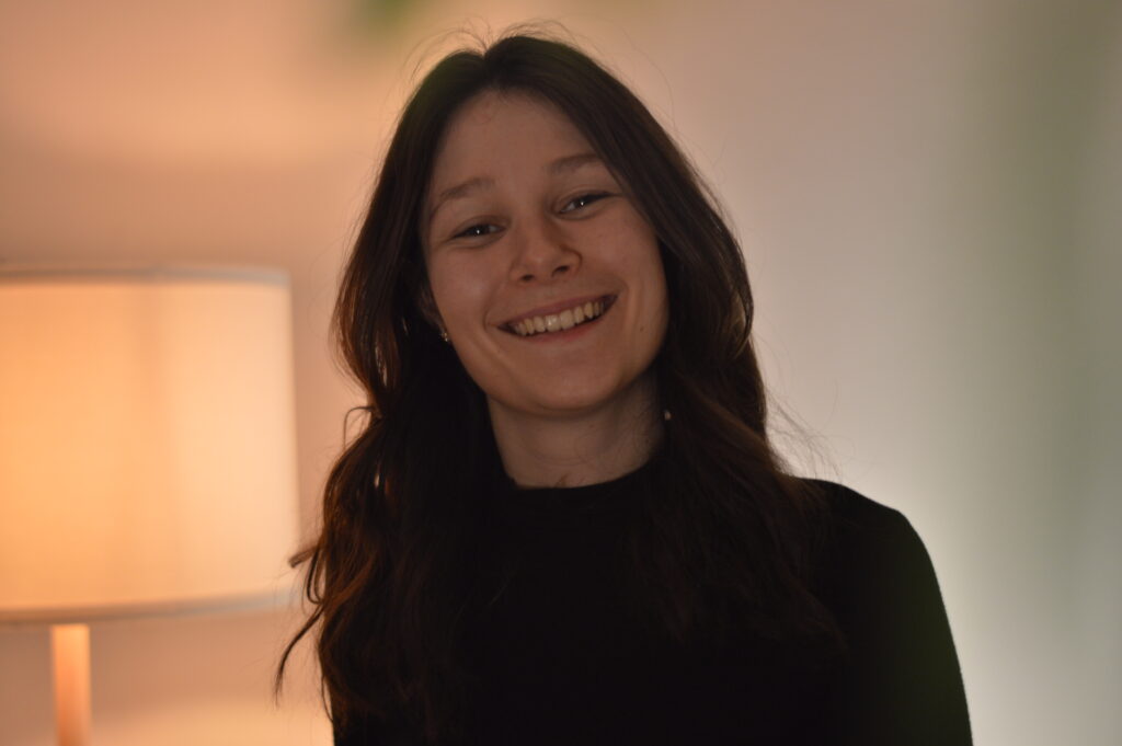 Eine junge Frau mit langen braunen Haaren, die ein schwarzes Oberteil trägt, lächelt warm in einem Innenraum mit einer brennenden Lampe im Hintergrund.