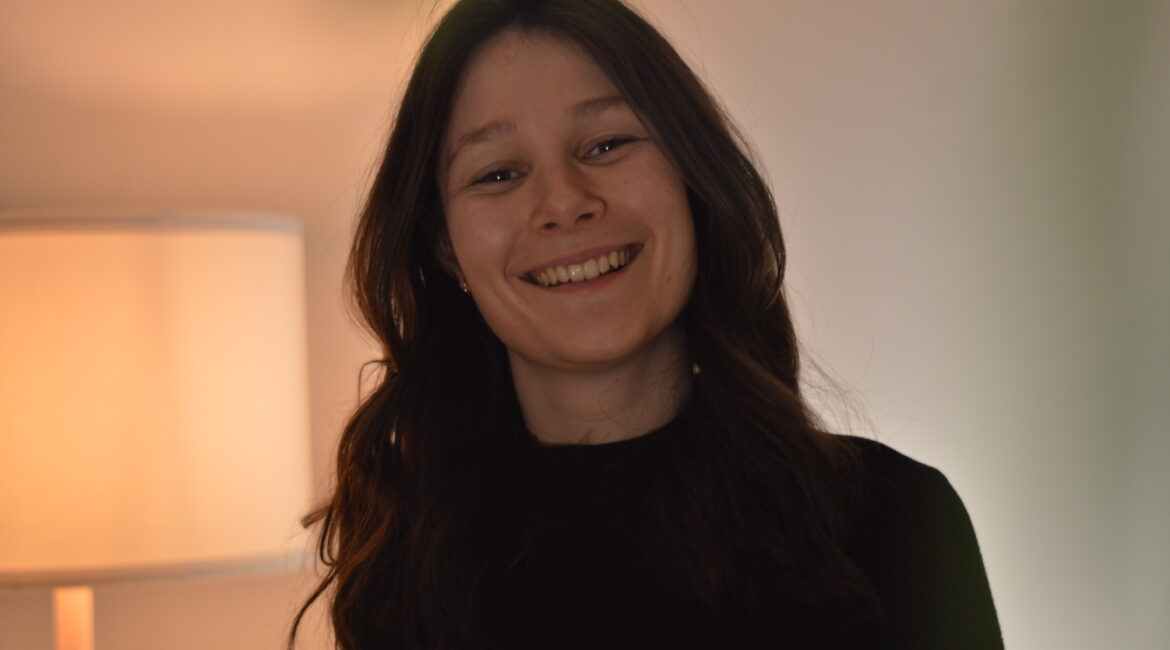 Eine junge Frau mit langen braunen Haaren, die ein schwarzes Oberteil trägt, lächelt warm in einem Innenraum mit einer brennenden Lampe im Hintergrund.