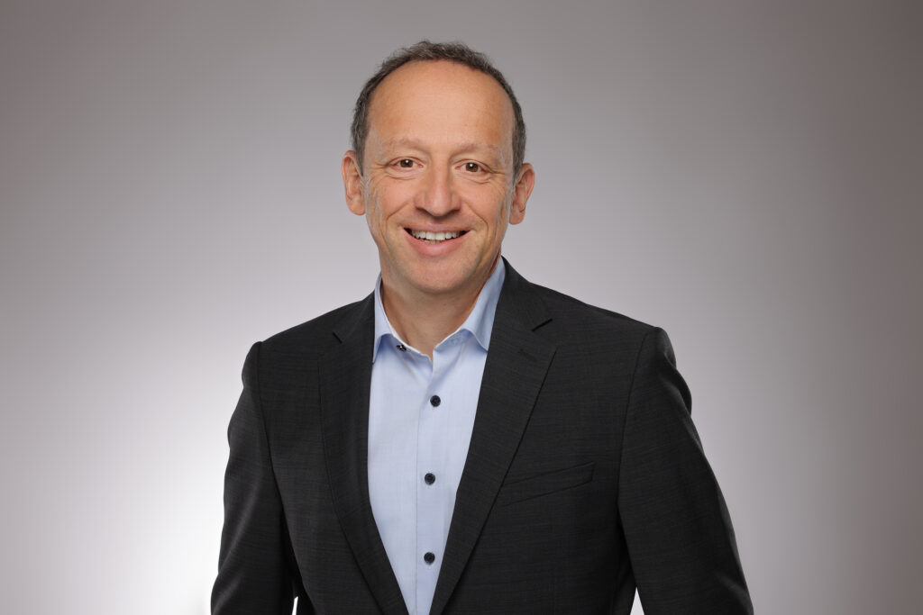 Ein Mann in einem dunklen Anzug und hellblauem Hemd lächelt vor einem schlichten, hellgrauen Hintergrund in die Kamera.