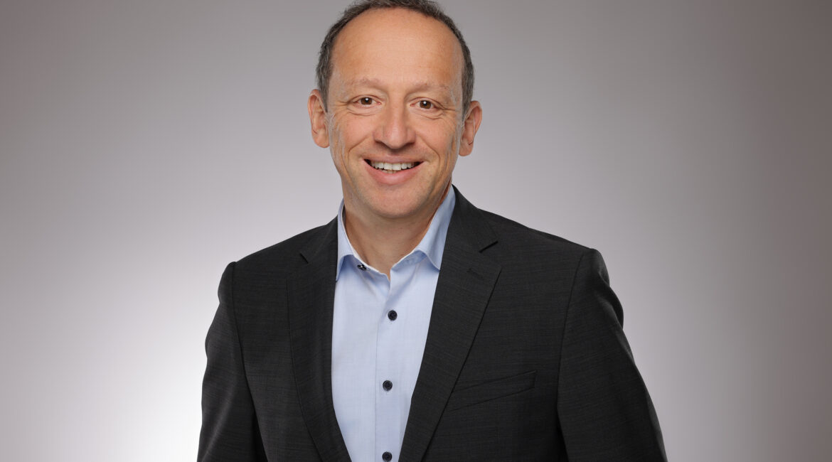 Ein Mann in einem dunklen Anzug und hellblauem Hemd lächelt vor einem schlichten, hellgrauen Hintergrund in die Kamera.