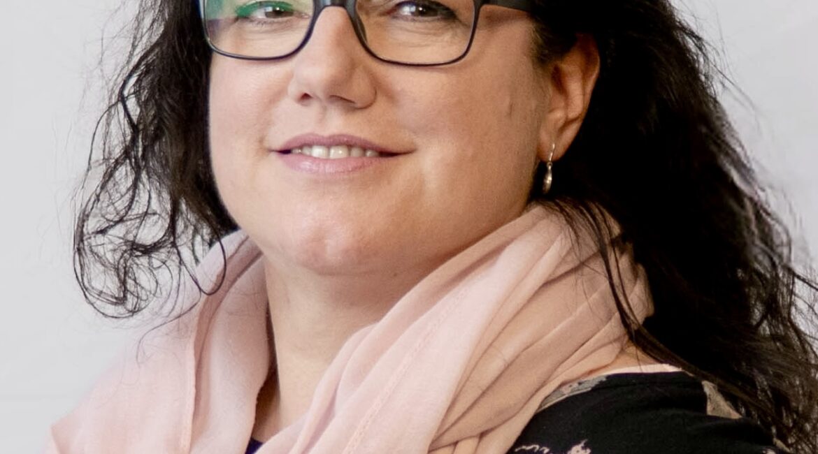 Eine Frau mit gewelltem dunklem Haar, Brille und einem blassrosa Schal lächelt leicht und trägt ein schwarzes Oberteil mit hellem Muster vor einem schlichten hellen Hintergrund.