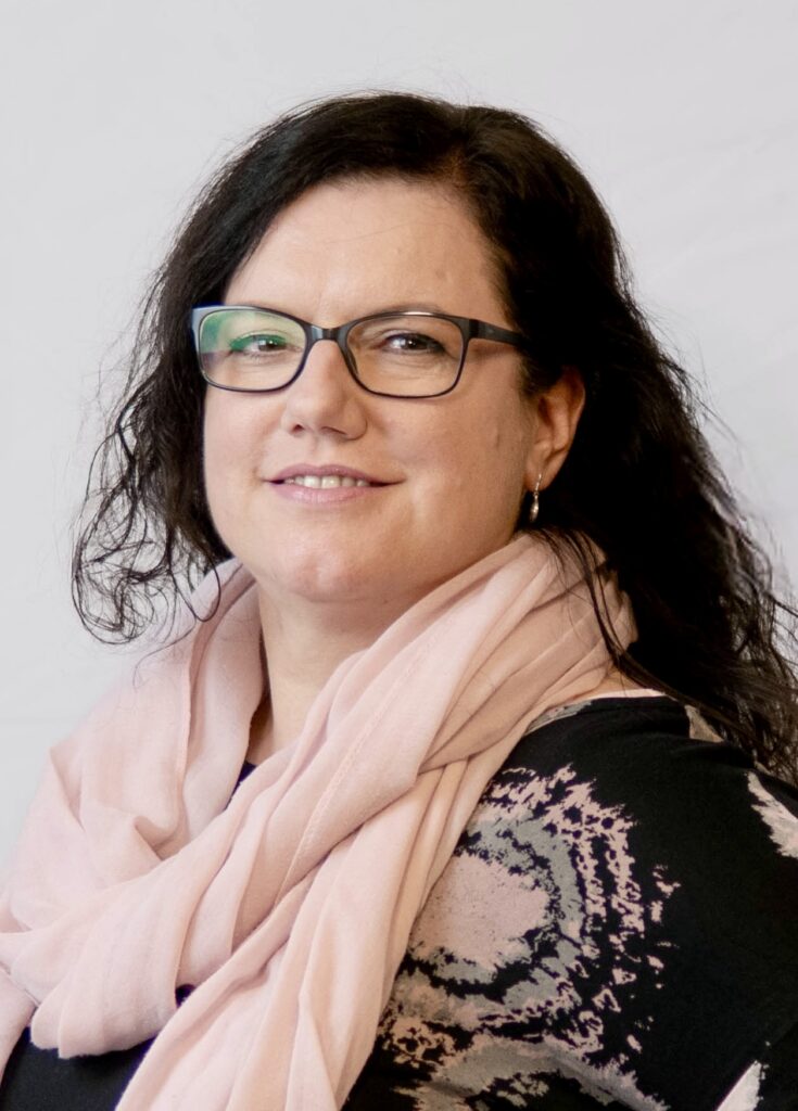 Eine Frau mit gewelltem dunklem Haar, Brille und einem blassrosa Schal lächelt leicht und trägt ein schwarzes Oberteil mit hellem Muster vor einem schlichten hellen Hintergrund.