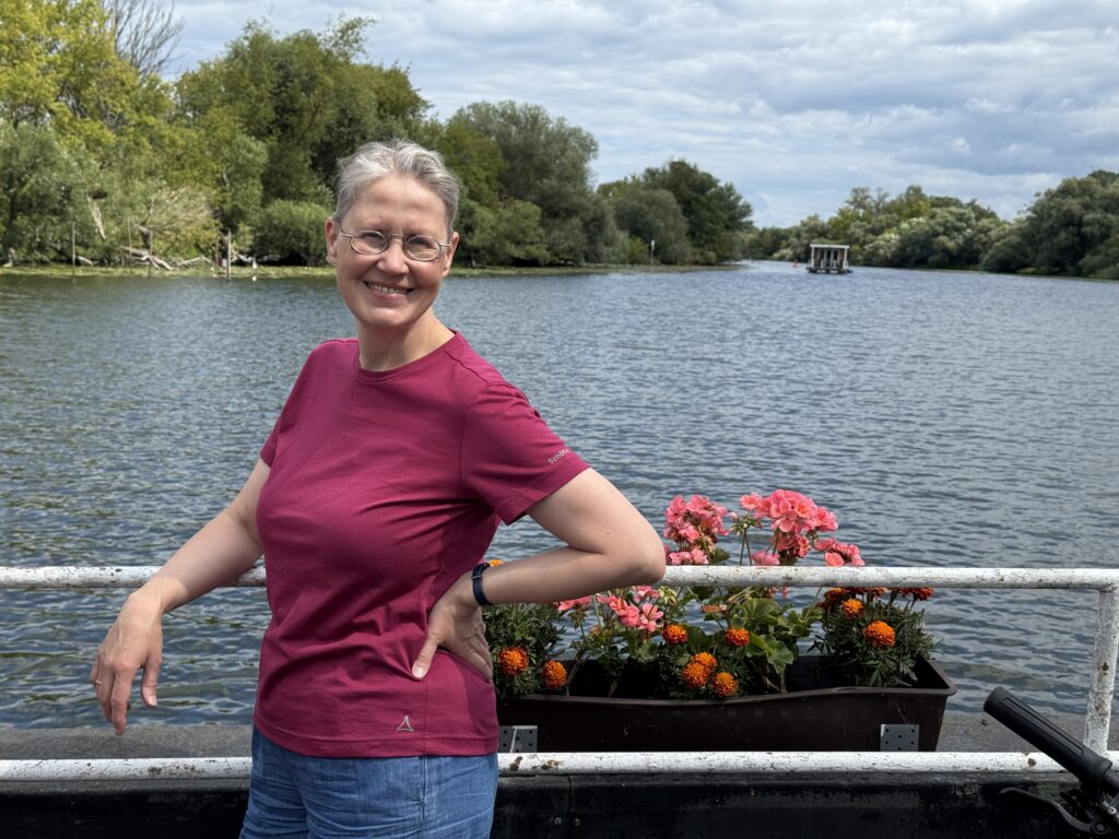 Eine lächelnde Person in einem magentafarbenen Hemd steht an einem Geländer am Flussufer mit bunten Blumen, Bäumen und einem bewölkten Himmel im Hintergrund.