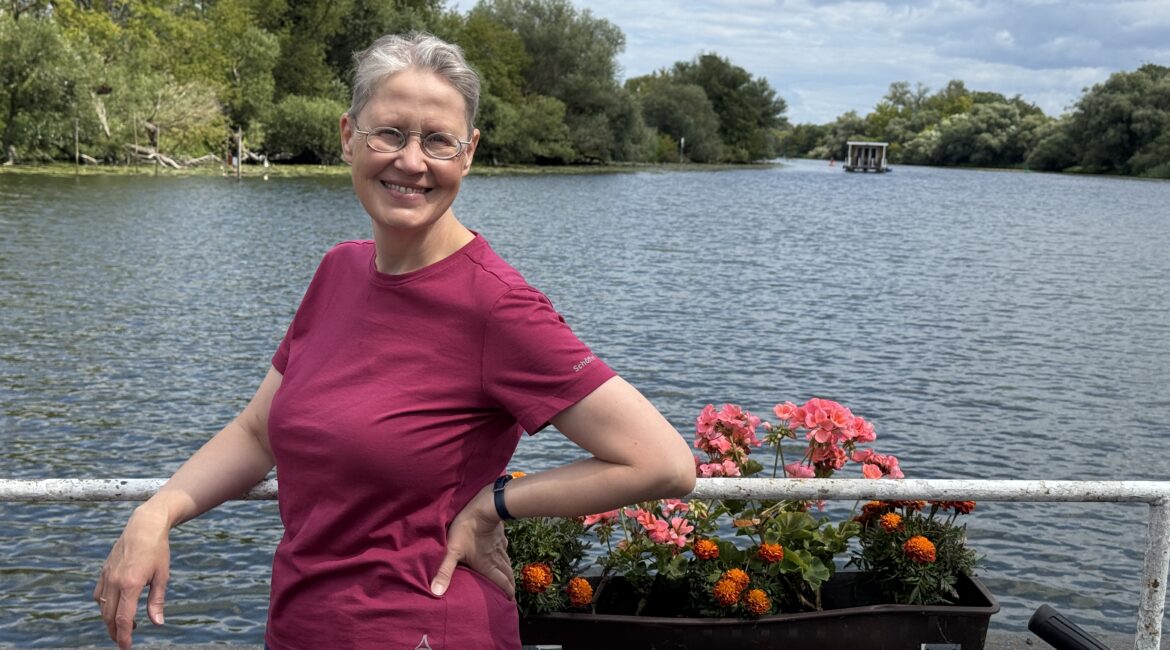 Eine lächelnde Person in einem magentafarbenen Hemd steht an einem Geländer am Flussufer mit bunten Blumen, Bäumen und einem bewölkten Himmel im Hintergrund.