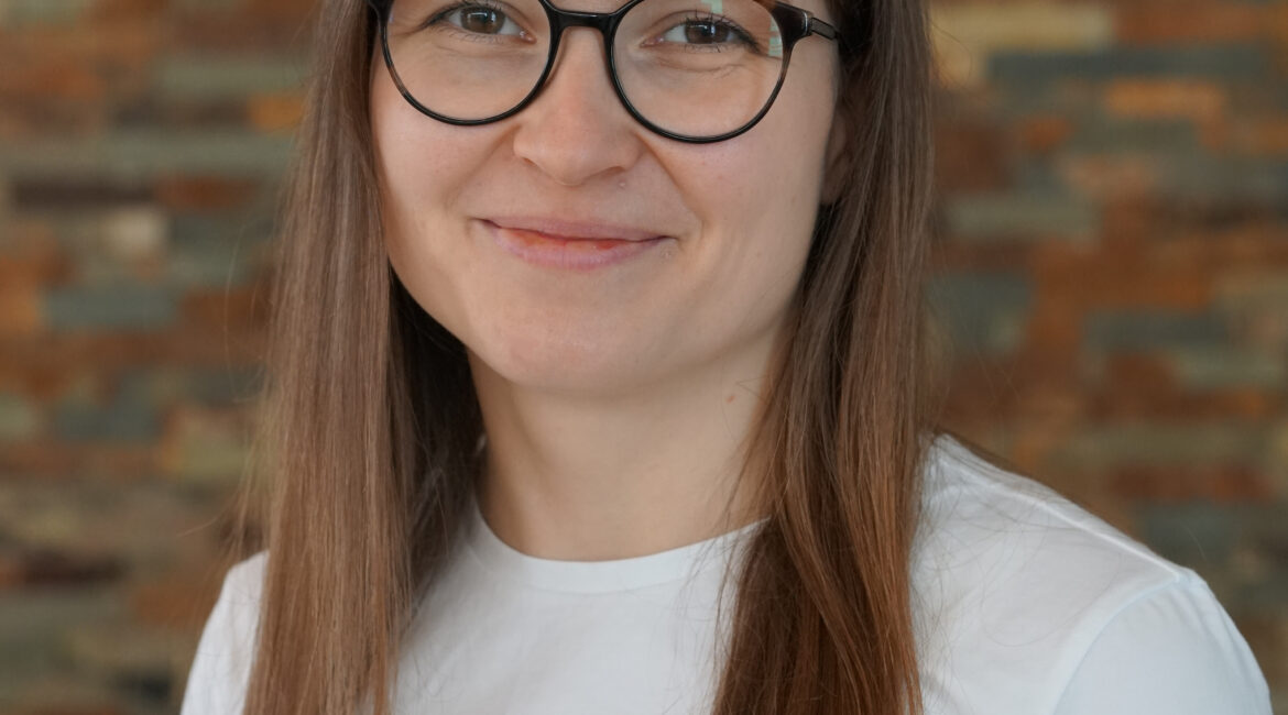 Eine Frau mit glattem braunem Haar und Brille, die ein weißes Oberteil trägt, lächelt sanft vor dem Hintergrund einer strukturierten Ziegelwand.