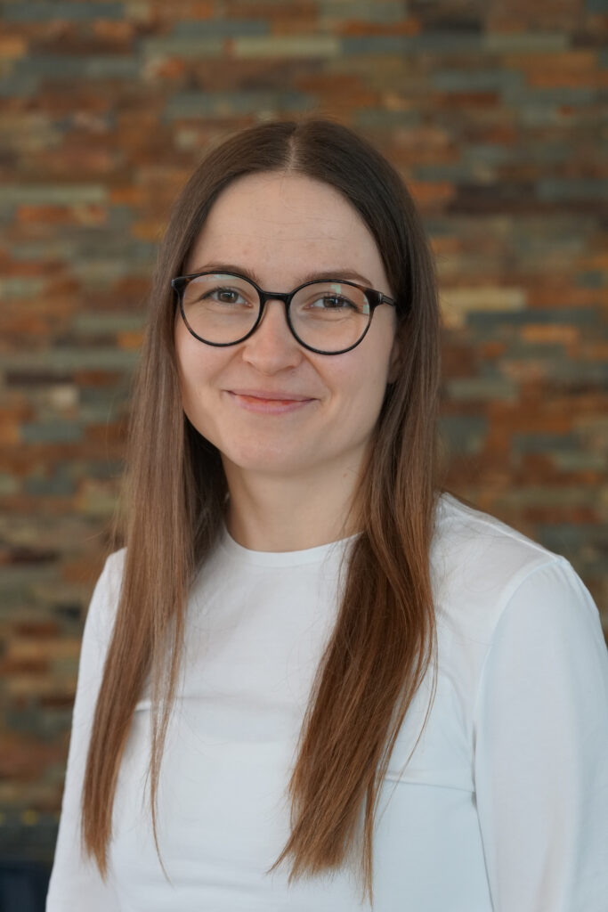 Eine Frau mit glattem braunem Haar und Brille, die ein weißes Oberteil trägt, lächelt sanft vor dem Hintergrund einer strukturierten Ziegelwand.
