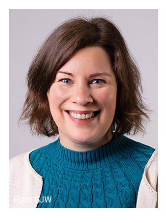Eine Frau mit kurzen braunen Haaren, die einen blaugrünen Pullover und eine helle Jacke trägt, lächelt in die Kamera vor einem schlichten Hintergrund. Foto: CJW steht in der unteren linken Ecke.
