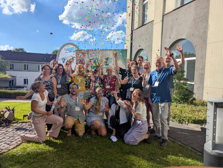 Eine Gruppe von Menschen, die im Freien feiern, bunte Konfetti in die Luft werfen und an einem sonnigen Tag vor einem Gebäude lächeln.