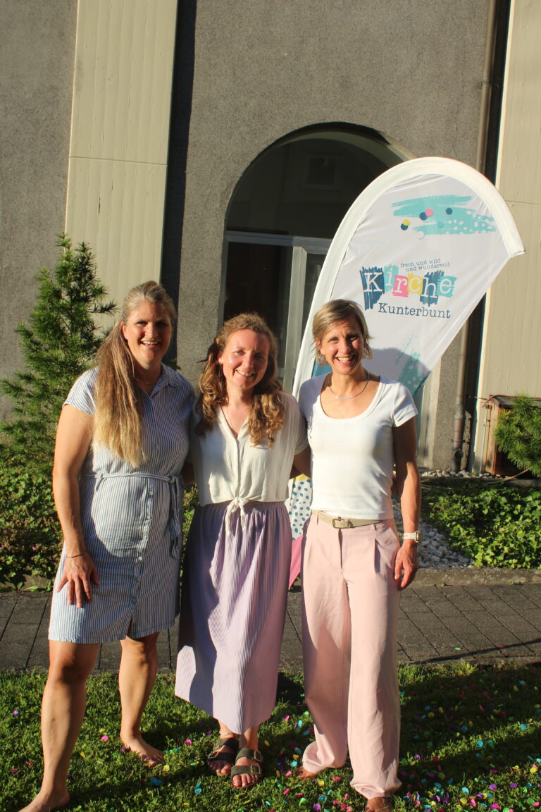 Drei Frauen stehen lächelnd zusammen draußen im Sonnenlicht, hinter ihnen sind Grünpflanzen und ein buntes Transparent mit der Aufschrift „Kirche Kunterbunt“ zu sehen.