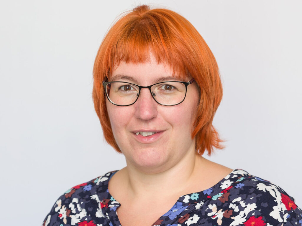 Eine Frau mit glattem, kinnlangem orangefarbenem Haar und Brille lächelt. Sie trägt ein marineblaues Oberteil mit einem bunten Blumenmuster. Der Hintergrund ist einfarbig hellgrau.