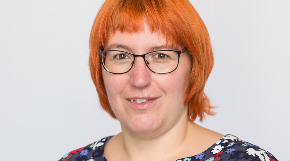 Eine Frau mit glattem, kinnlangem orangefarbenem Haar und Brille lächelt. Sie trägt ein marineblaues Oberteil mit einem bunten Blumenmuster. Der Hintergrund ist einfarbig hellgrau.