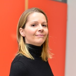 Eine Frau mit glattem blondem Haar und schwarzem Rollkragenpullover steht vor einem orange-beigen Hintergrund und schaut selbstbewusst in die Kamera.
