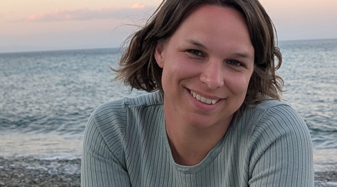 Eine Frau mit kurzen braunen Haaren sitzt bei Sonnenuntergang an einem felsigen Strand und lächelt, im Hintergrund sind das Meer und der pastellfarbene Himmel zu sehen.