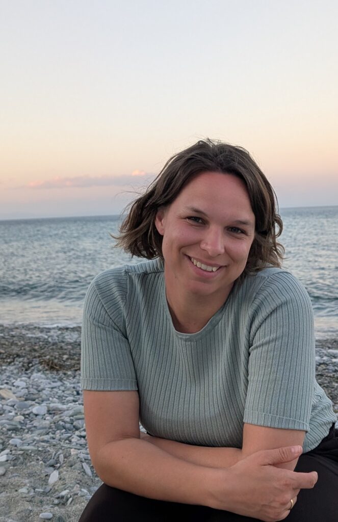 Eine Frau mit kurzen braunen Haaren sitzt bei Sonnenuntergang an einem felsigen Strand und lächelt, im Hintergrund sind das Meer und der pastellfarbene Himmel zu sehen.