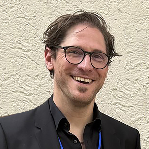Ein Mann mit Brille und dunklen Haaren, der lächelt, eine schwarze Jacke und ein blaues Umhängeband trägt und vor einer leicht strukturierten Wand steht.