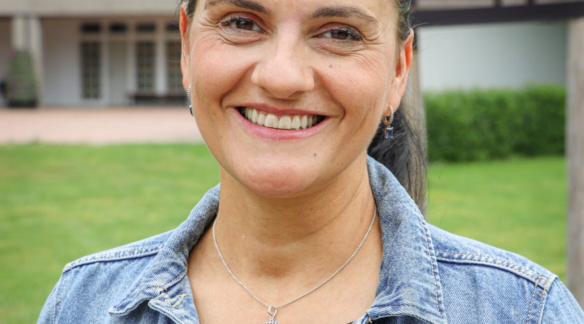 Eine lächelnde Frau mit nach hinten gebundenen Haaren trägt eine Jeansjacke und eine Halskette mit einem Kreuz.